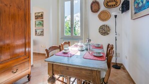 Un comedor con una mesa de madera, sillas y un ventanal con cortinas. Hay cestas colgadas en la pared y un cuadro enmarcado.