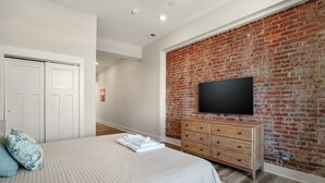 Un dormitorio con pared de ladrillo, un televisor de pantalla plana, un aparador de madera y una cama con almohadas.