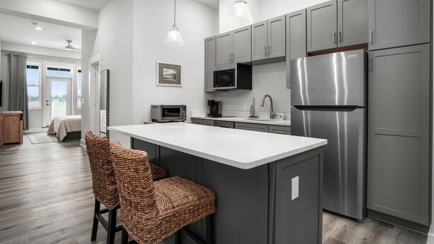 A modern kitchen with grey cabinets, a stainless steel refrigerator, and a central island with wicker chairs.