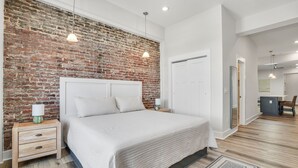 A bedroom with a brick wall, a bed with a white headboard, bedside tables with lamps, and a wooden floor.