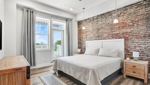 A bedroom with a brick accent wall, a large bed, a nightstand with a lamp, and a view of the outdoors through a window.
