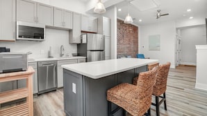 A modern kitchen with grey cabinets, stainless steel appliances, a central island, wicker bar stools, and a brick accent wall.
