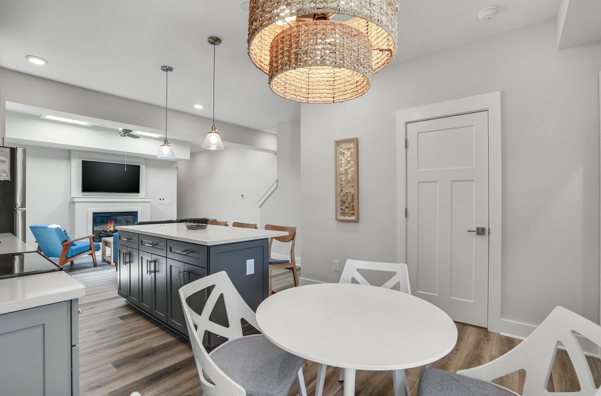 A modern kitchen with a central island, white dining table, and chairs. There is a flat-screen TV mounted above the island, and a large pendant light hangs above the dining area.