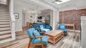 Een moderne woonkamer met een bank, stoelen en een salontafel. Achtergrond is een keuken met een koelkast en een bakstenen muur.