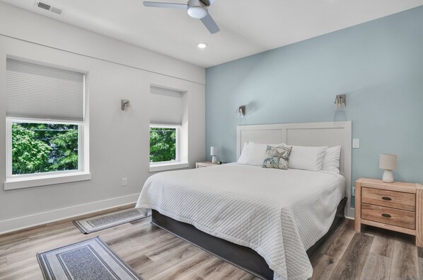 A bedroom with a large bed, two bedside tables, a ceiling fan, and two windows with blinds.
