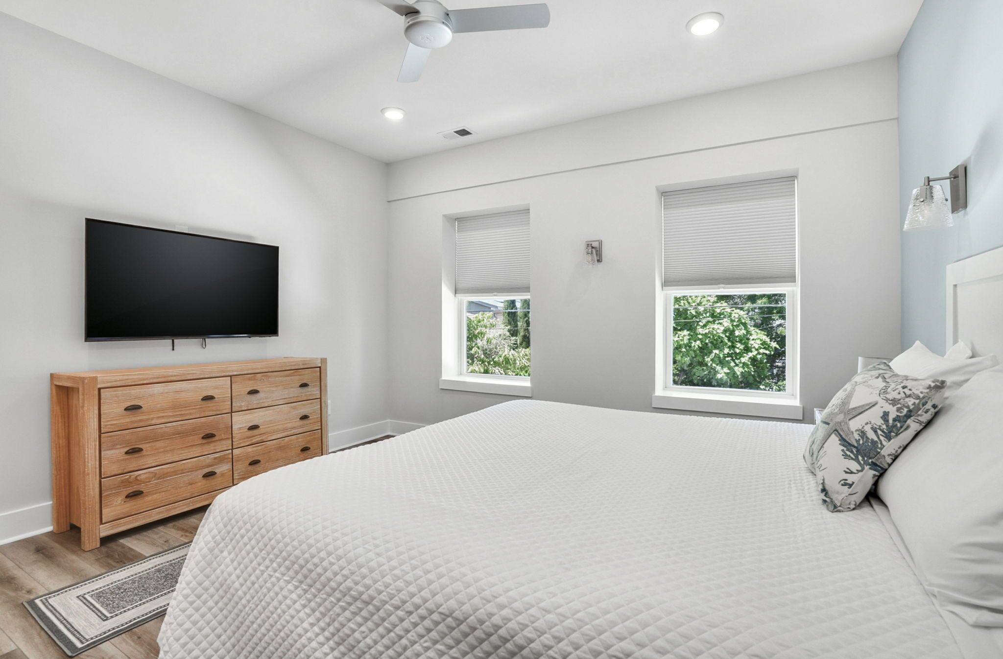 A bedroom with a bed, a TV, a dresser, and two windows with blinds.