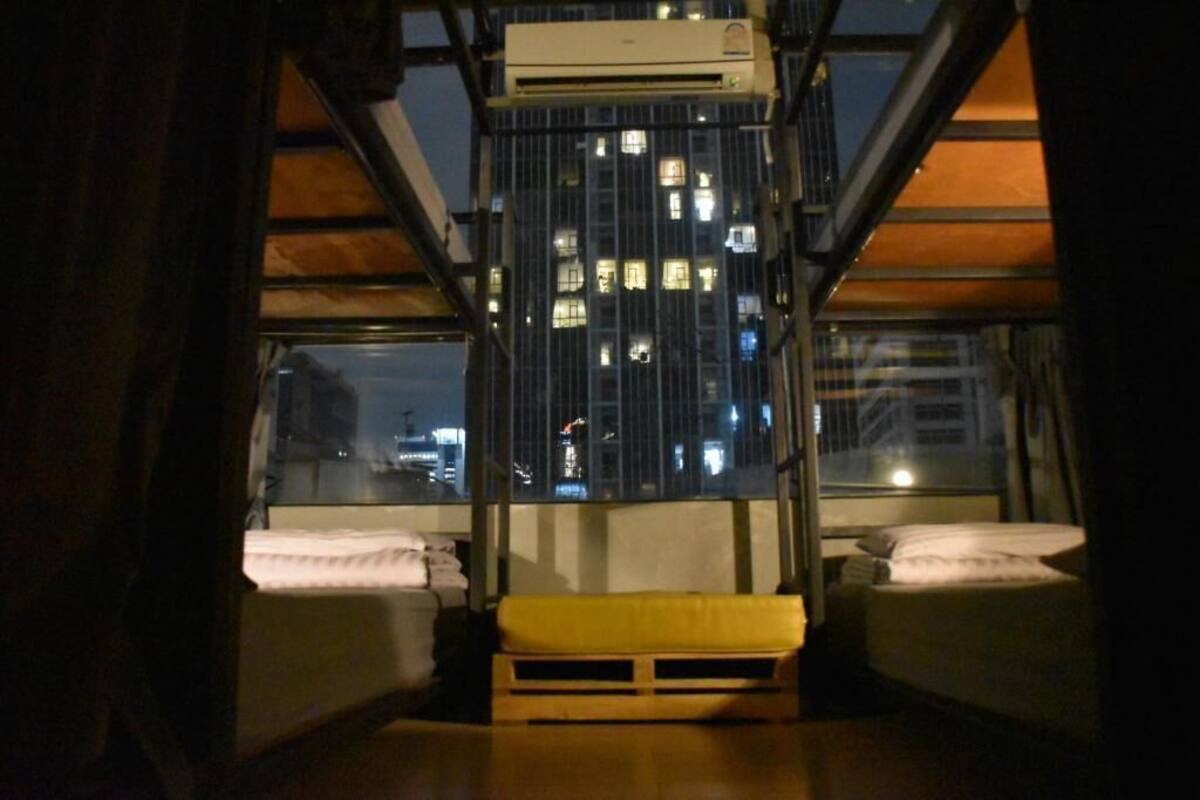 A room with two beds, a yellow bench, and a window overlooking a cityscape at night.