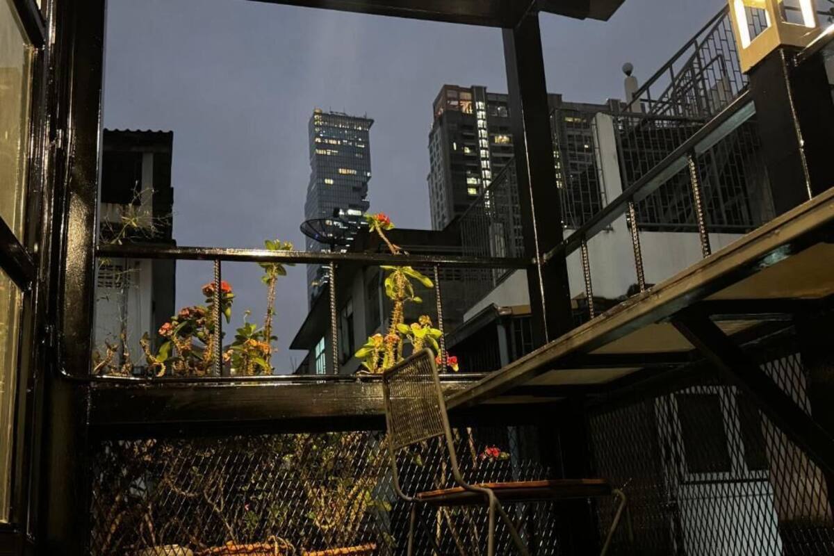 A balcony with potted plants and a chair, overlooking a cityscape with tall buildings.
