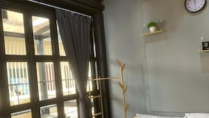 A bedroom with a bed, a clock, a plant on a shelf, and a window with curtains. - STAY AT PAN (Bangkok)