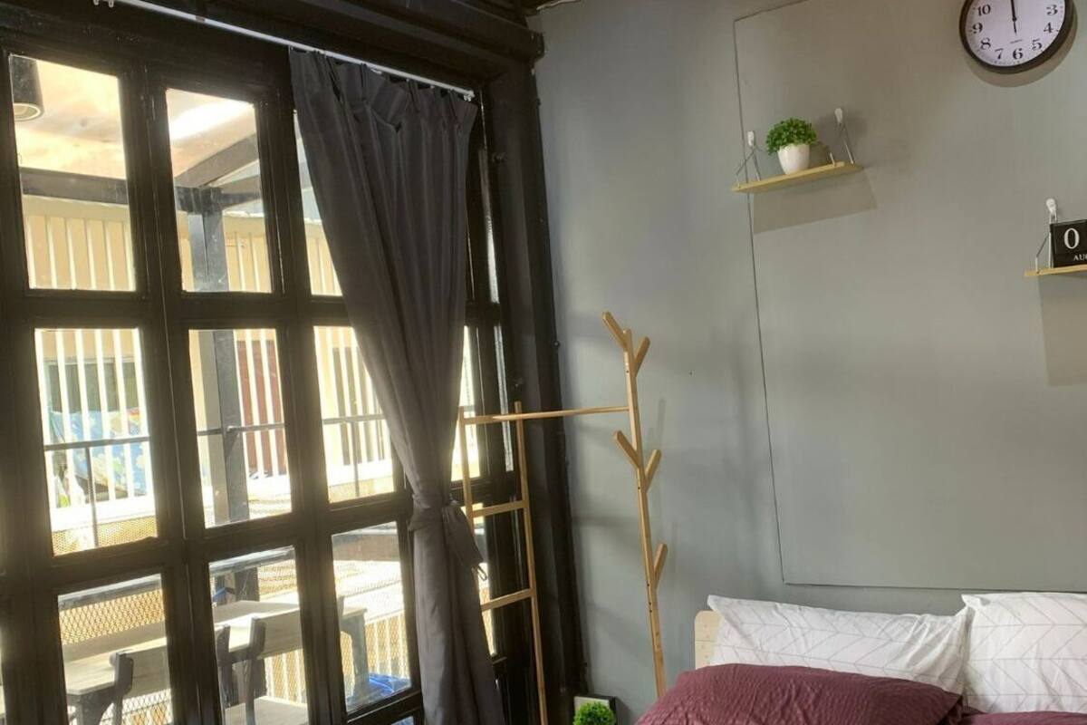 A bedroom with a bed, a clock, a plant on a shelf, and a window with curtains.