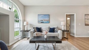 A modern living room with a sofa, coffee table, and lamps. There is a painting on the wall and a window with a view. - Casa de Palmera - Team Joseph Ellen (Palm Coast)