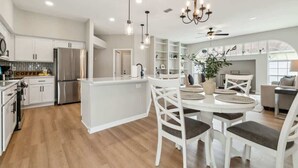 A modern kitchen with white cabinetry, a central island, and a dining area with white chairs. - Casa de Palmera - Team Joseph Ellen (Palm Coast)