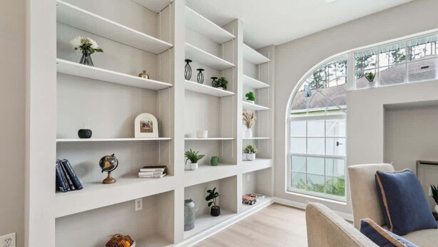 A modern living room with white shelving units, a sofa with blue cushions, and a large window with a view of greenery. - Casa de Palmera - Team Joseph Ellen (Palm Coast)