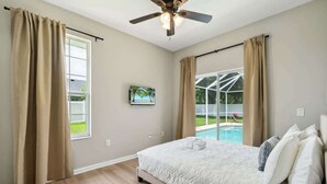 A bedroom with a bed, a ceiling fan, a window with curtains, a painting, and a view of a swimming pool. - Casa de Palmera - Team Joseph Ellen (Palm Coast)
