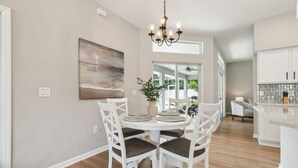 A dining area with a round table, white chairs, a chandelier, and a painting on the wall. - Casa de Palmera - Team Joseph Ellen (Palm Coast)
