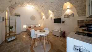 Una habitación compacta con techo de piedra, una cama con colcha color beige, una pequeña mesa redonda con sillas, un televisor blanco en la pared y una zona de kitchenette.