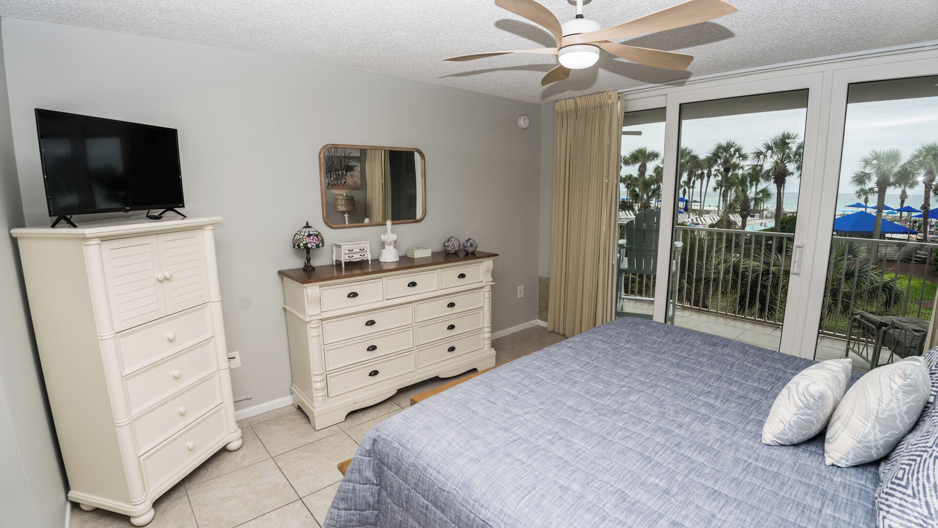 A bedroom with a bed, dresser, television, and a balcony with a view of palm trees and a beach.