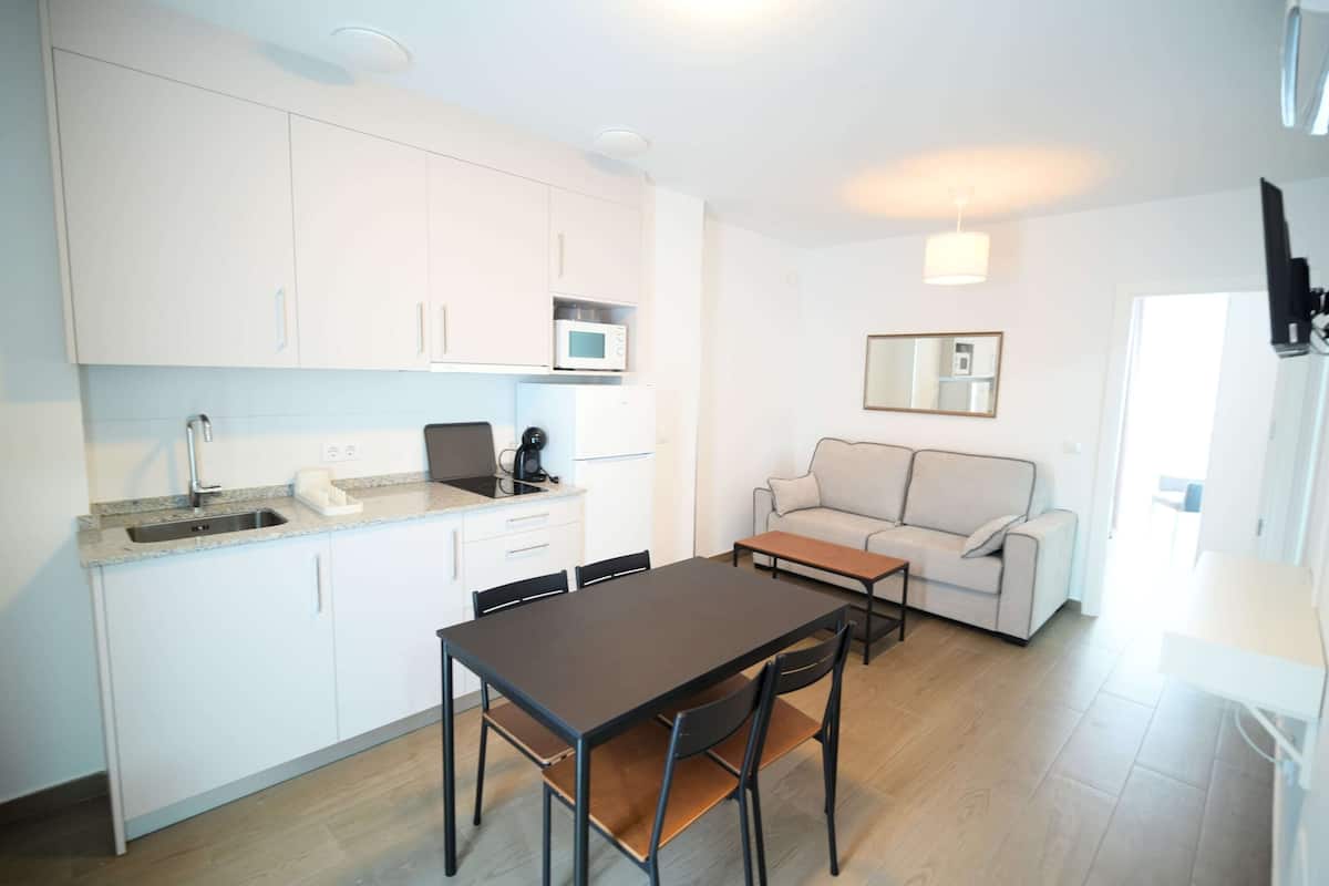 A compact kitchen with white cabinets, a microwave, and a coffee maker. There is a dining table with chairs and a sofa in the living area.