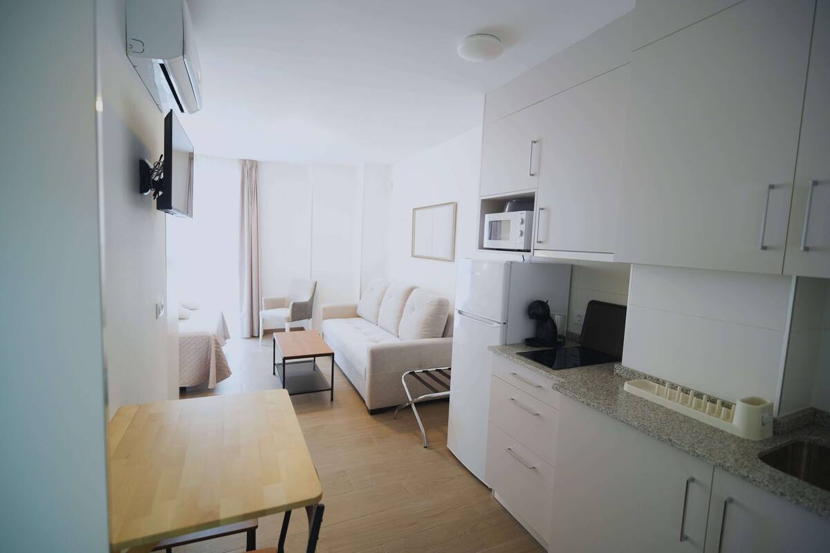 A compact kitchen with white cabinets, a microwave, and a sink. There is a small dining table and a sofa in the adjacent living area.