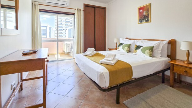 A hotel room with a bed, a desk, a chair, a window with curtains, an air conditioning unit, and a painting on the wall.