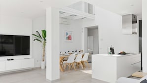 Un salon moderne doté d’un téléviseur à écran plat, d’un coin repas avec une table et des chaises en bois, et d’une cuisine avec un plan de travail et un évier blancs.