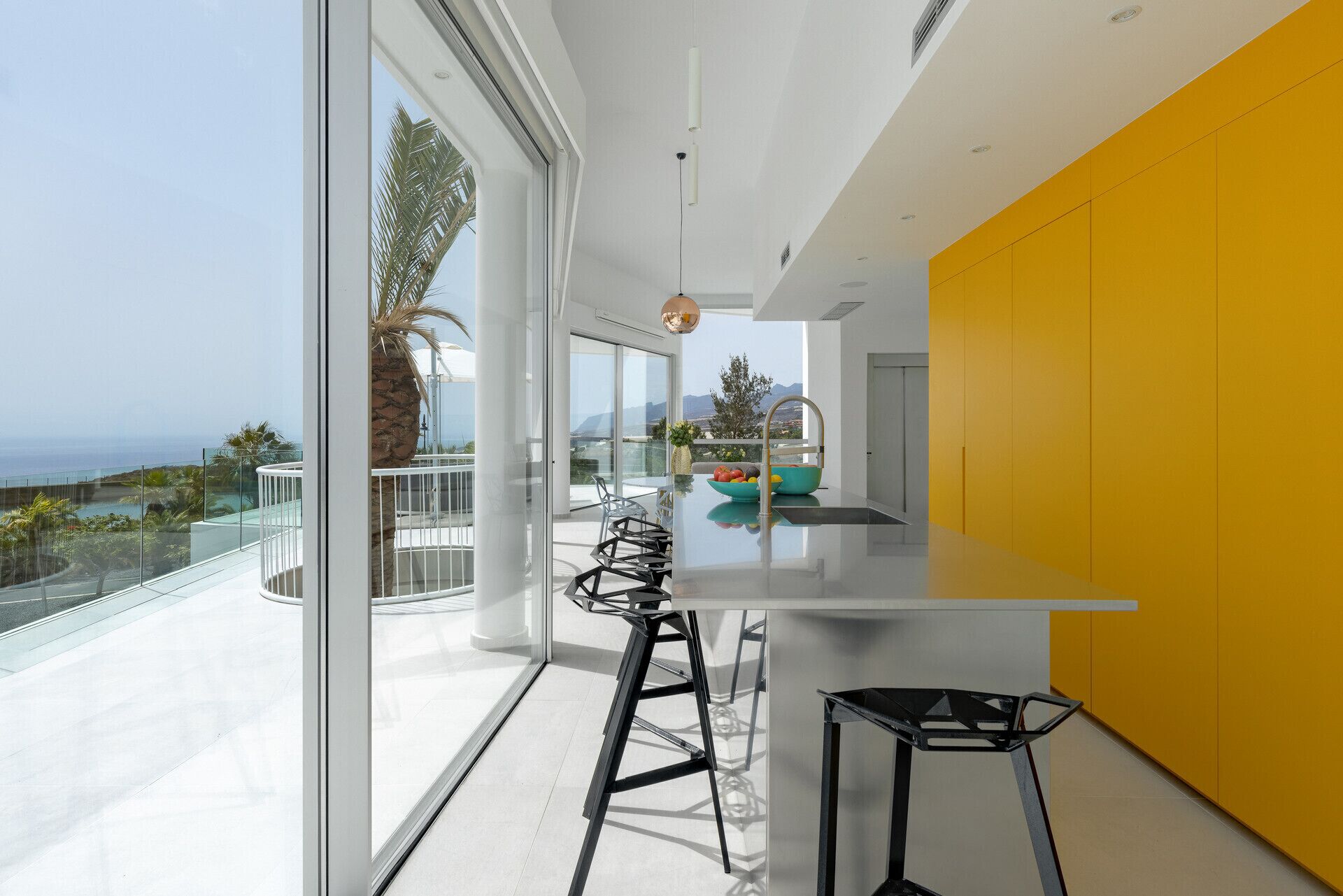 Une cuisine moderne dotée d’un grand îlot central, d’un mur d’accent jaune et offrant une vue sur l’océan.