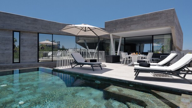 A modern outdoor pool area with lounge chairs and umbrellas, large windows, and a clear view of the surrounding landscape. - Villa Origen Exclusive (Adeje)