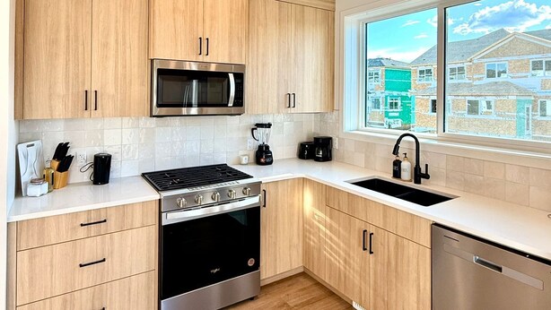 A modern kitchen with wooden cabinets, stainless steel appliances, a dishwasher, and a window with a view of houses.