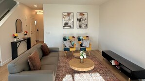 A modern living room with a grey sofa, two armchairs, a coffee table, and a black bench. There are two abstract paintings on the wall, a staircase, and a small table with flowers.