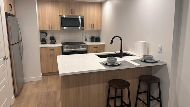 Una cocina moderna con gabinetes de madera, estufa de acero inoxidable, microondas y un lavamanos. Hay dos taburetes en la isla.