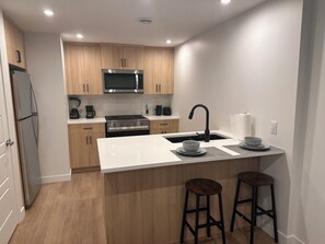 Una cocina moderna con gabinetes de madera, estufa de acero inoxidable, microondas y un lavamanos. Hay dos taburetes en la isla.