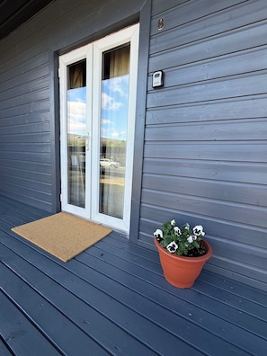 Une maison à l’extérieur gris, avec une porte-fenêtre coulissante, un paillasson marron et un pot de fleurs contenant des plantes aux fleurs blanches et violettes.