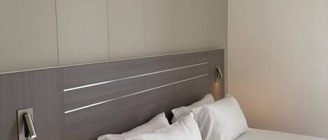 A hotel room with a bed, a bedside table, a wall-mounted light, and a power outlet.