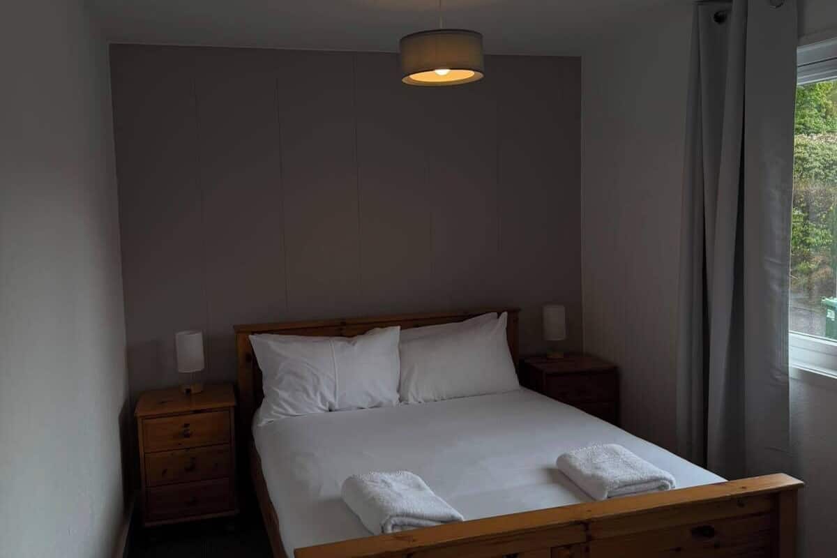 Une chambre d’hôtel avec un lit en bois, du linge de lit blanc, une table de chevet avec une lampe et une fenêtre avec des rideaux.