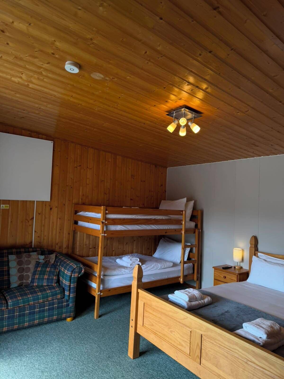A bunk bed room with a wooden ceiling, a small bedside table with a lamp, and a plaid sofa.