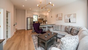 A modern living room with a sofa, coffee table, and purple armchair. A kitchen with a refrigerator and bar stools is visible in the background. - Turf ROOFTOP + Sleeps Up to 16 If Renting Multiple Units! Unit B (Wilmington)