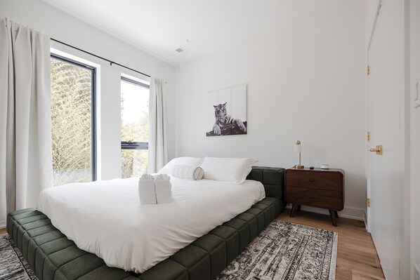 A bedroom with a large bed, a nightstand, a framed picture on the wall, and a window with curtains.