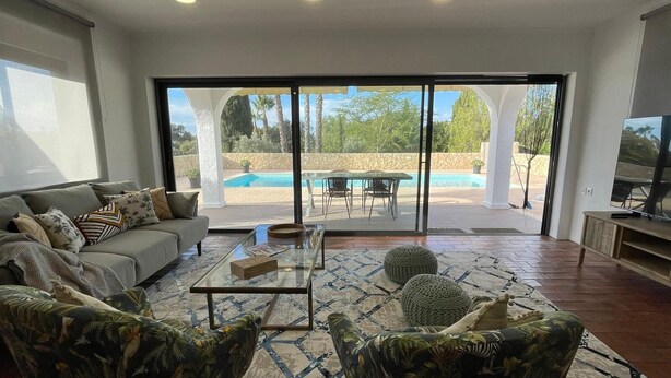 Un salon spacieux comprenant un canapé, deux fauteuils, une table basse et un téléviseur. Une grande porte-fenêtre coulissante donne accès à un espace extérieur avec une piscine et du mobilier de jardin.