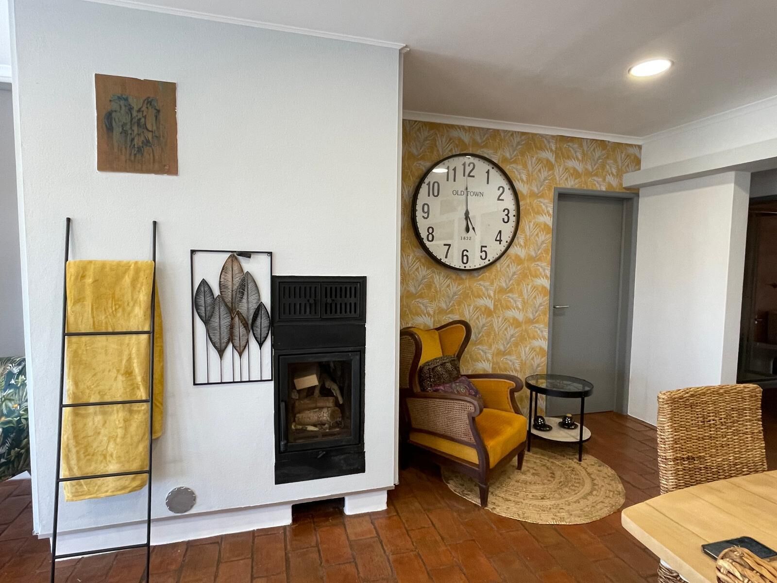 Una sala de estar con chimenea, un reloj de pared grande, un sillón amarillo y una silla de mimbre.