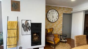 Una sala de estar con chimenea, un reloj de pared grande, un sillón amarillo y una silla de mimbre.