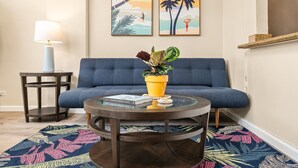 A living room with a blue sofa, a glass coffee table with books, a potted plant, and a patterned rug.
