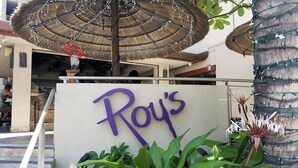 A hotel entrance with a sign reading "Roy's," surrounded by lush greenery and a thatched umbrella.