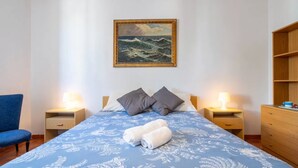 A hotel room with a bed, two bedside tables with lamps, a blue patterned bedspread, two pillows, a blue chair, and a wooden dresser. - Large seaside apartment with patio (Lu Bagnu)