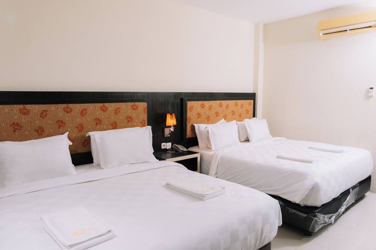 Habitación de hotel con dos camas, ropa de cama blanca, una mesita de noche con lámpara y aire acondicionado instalado en la pared.