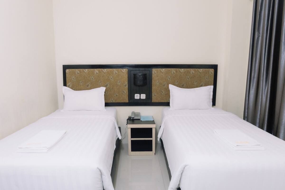 Habitación de hotel con dos camas individuales, una mesita de noche pequeña con teléfono y cabecera con panel decorativo.