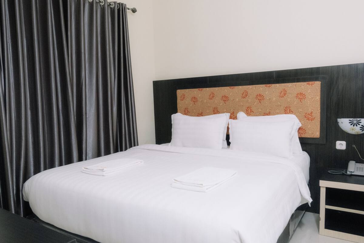 Una cama bien hecha con sábanas y toallas blancas, cabecera oscura con estampado floral, mesita de noche con teléfono y una cortina en la ventana.