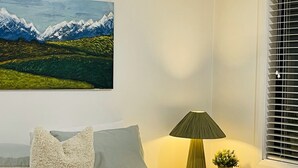 Makuuhuone, jossa on sänky, tyynyjä, yöpöytä lampun kanssa, vuoristomaisemaa esittävä taulu ja ikkuna, jossa on sälekaihtimet.