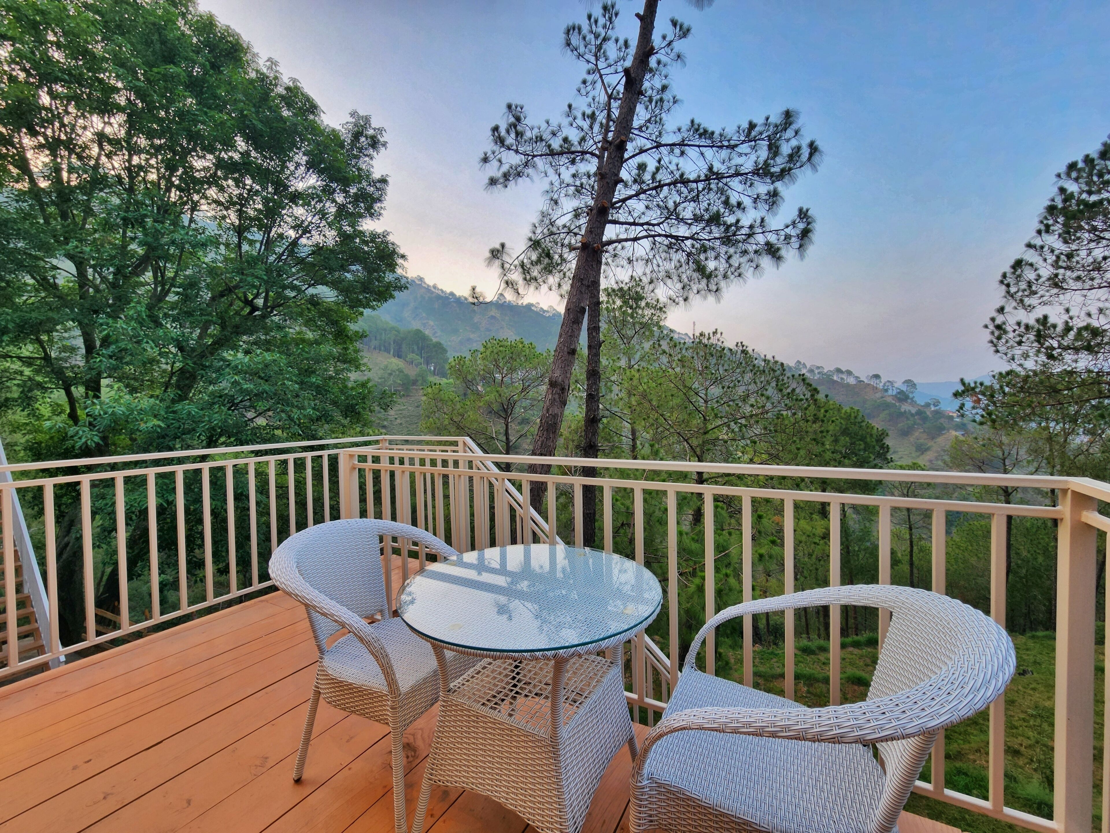 A wooden deck with wicker furniture and a glass table, overlooking a forested landscape with distant hills.