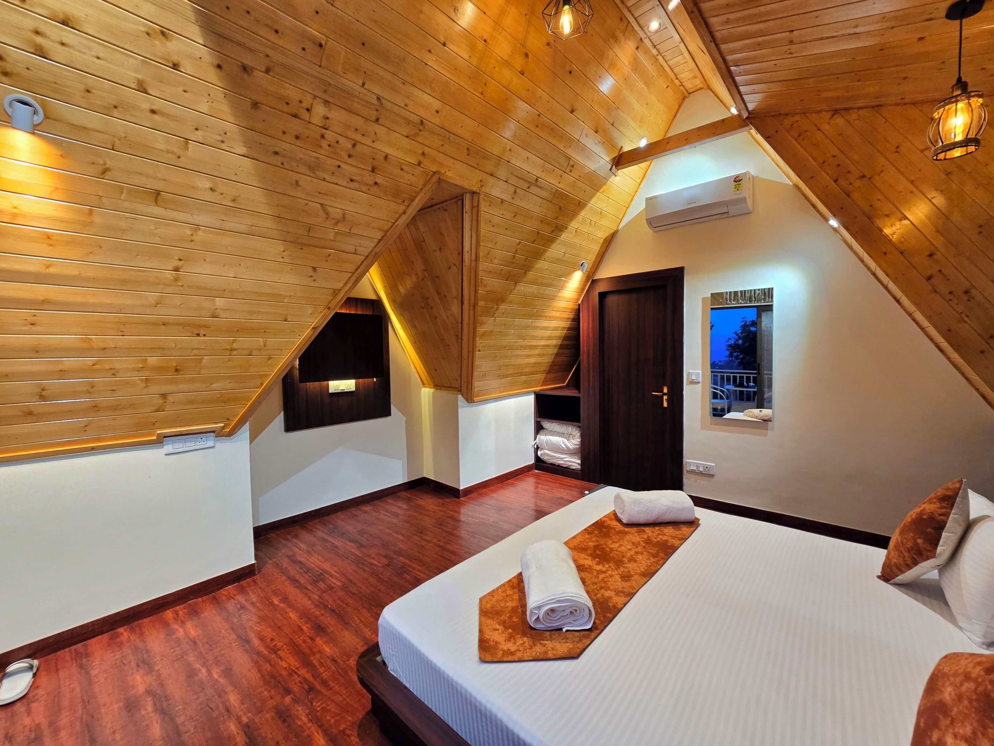 A bedroom with a sloped ceiling, wooden walls, a bed with a white mattress and brown headboard, a window with a view, and a built-in shelf with towels.