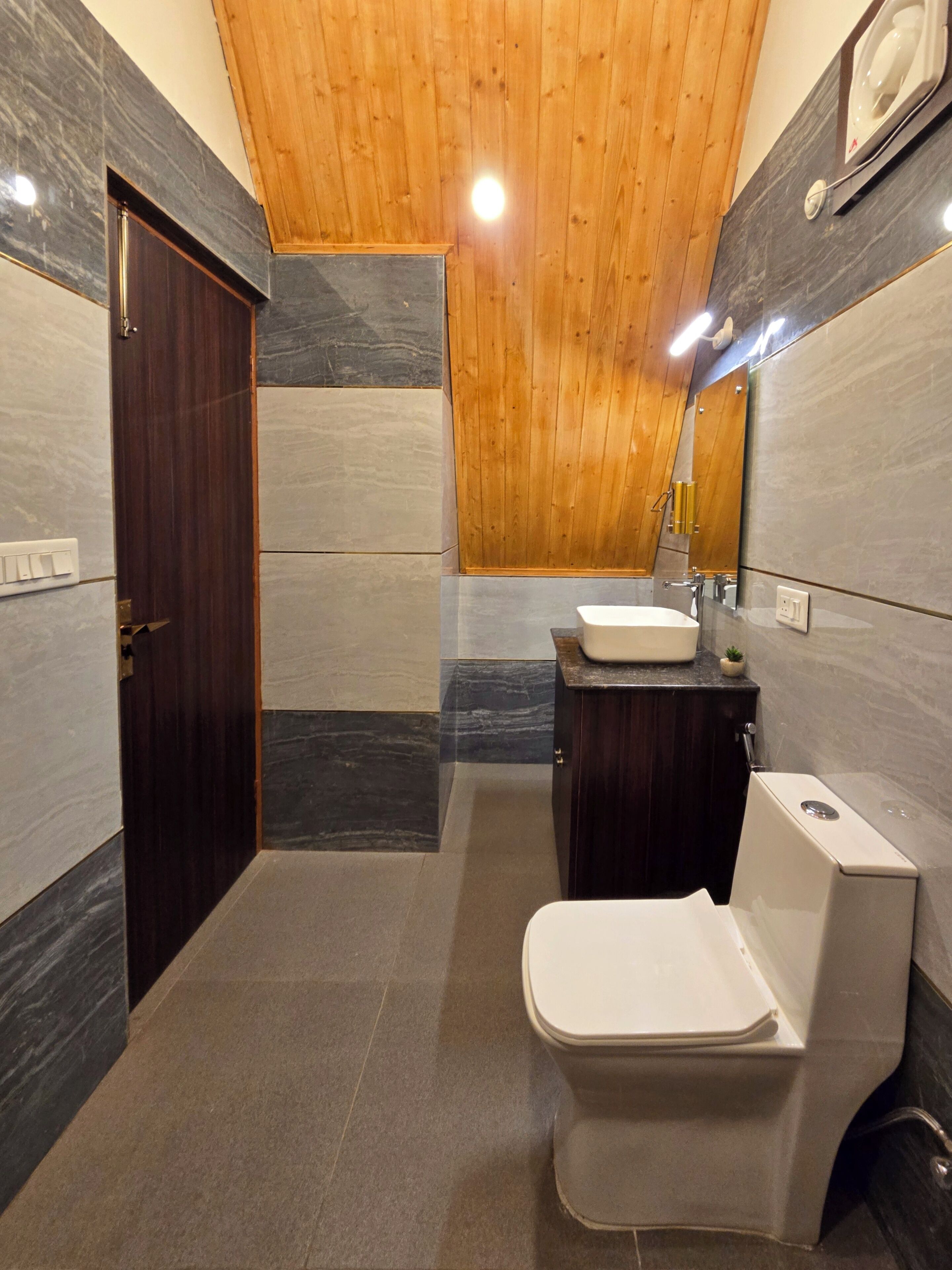 A modern bathroom with a wooden ceiling, a marble-tiled floor, a wooden door, a white toilet, a sink, and a mirror.